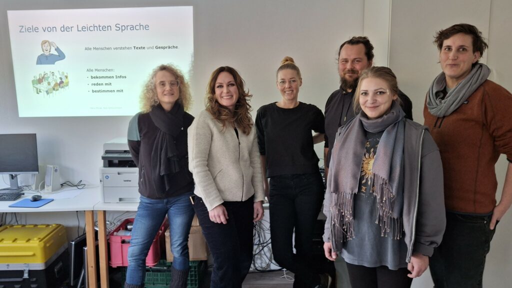 Auf dem Bild ist eine Gruppe Menschen zu sehen, im Hintergrund an der Wand ein Präsentationsslide zu Leichter Sprache.