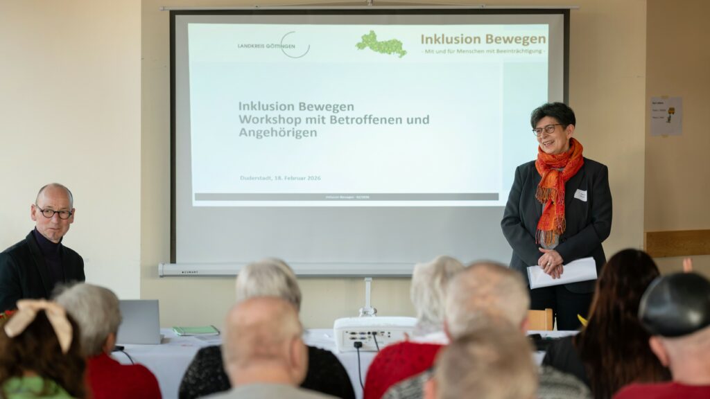 Das Bild zeigt eine Gruppe Menschen vor einer Leinwand, es steht rechts Regina Meyer und begrüßt die Menschen.