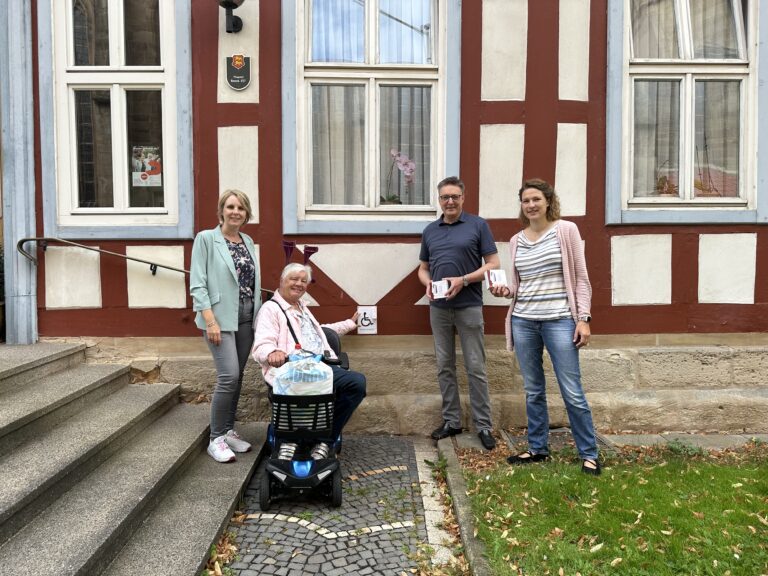 Vier Personen vor dem Pfarrbüro. Eine Frau auf einem Elektromobil zeigt auf die Funkklingel neben der Treppe. Zwei Personen halten Klingelverpackungen.